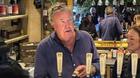 PA Media/Neil Robinson Jeremy Clarkson inside his pub, in front of a big mirror, pulling a pint. A large throng of customers can be seen in the mirror.