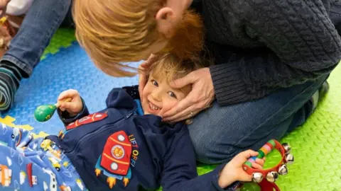 Chris Bailey A young child is lying on their dad's lap. The child is laughing and holding am instrument in each hand.
