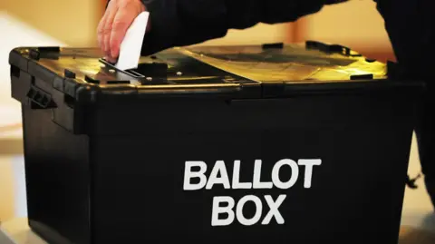 PA Media Someone is putting a white piece of paper into a black box that says 'ballot box' on the front in white writing. 