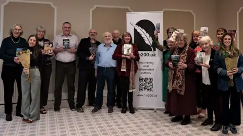 Colin Wade Colin Wade (fourth from left to right, holding a book) and Laury Conn (sixth from left to right wearing a blue shirt) on the launch of their 1in200 initiative at the Cotswold Lodge Hotel on Banbury Road in Oxford. They are surrounded by other people holding books and flowers. There is a white banner with the scheme's name, logo and QD code in the middle of the group. Everyone is smiling for the camera.