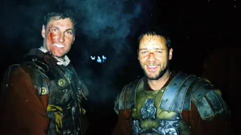 Andy Bowden Andy Bowden standing next to Russell Crowe while filming in Bourne Woods in 1999. Both are smiling and dressed in Roman armour costumes, with fake blood and bruising on their faces.