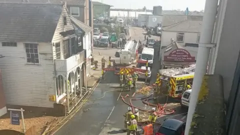 Martyn Bignell A white building on the left has fire damage that has caused the second floor to go black. On the road in front of the house are multiple firemen and a fire engine. 