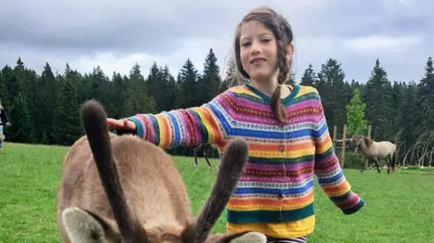 Family photo Aisha Cox smiles with a donkey. She is wearing a striped, multi coloured cardigan. In the background there is a forest lined with trees in a rural area.