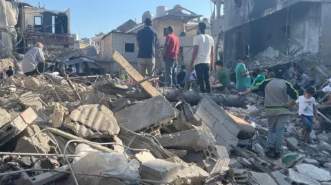 Aakriti Thapar/BBC Groups of men stand on a mound of rubble and twisted metal after an Israeli airstrike hit a building. 