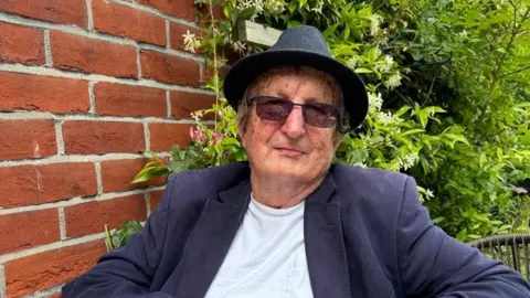 Contributed Andrew Lines smiles at the camera while sitting in a garden. He wears a black bowler hat, sunglasses, a navy suit jacket and white T-shirt. 