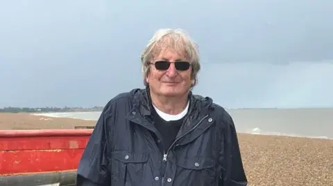Contributed Mr Lines smiles at the camera while standing on a beach. He has grey hair that rests over his forehead and wears sunglasses, a navy waterproof coat, black jumper and white T-shirt. A small red boat resting on the beach can be seen behind him. 