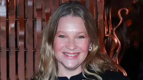 Joanna Page has blonde hair and blue eyes and is smiling at a camera. She is stood behind a bronze metallic background and is wearing a black top with gold detail