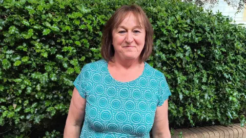 BBC Tricia Timms wearing a blue and white patterned top and standing in front of a holly bush. She has shoulder-length brown hair with a slight fringe, and is smiling at the camera with her arms down beside her.