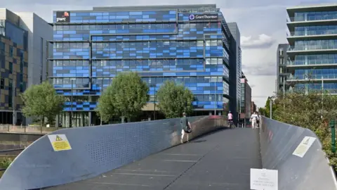 Google A screenshot from Google street view showing the metal bridge with thousands of small holes on either walled side. There are pedestrians and one cyclist crossing the bridge. In the background there is a large office building with lots of windows and blue panelling. 