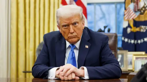 Getty Images Donald Trump sits at the desk in the Oval Office. He is wearing a navy suit jacket, a white collared shirt and a blue tie. There is a US flag pin on his lapel.