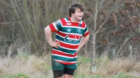 Submitted A teenager with brown windswept hair wearing a stripy green, red and white shirt with his hand on his hips and looking into the distance. He is also wearing green and black shorts and a woodland can be seen behind him.
