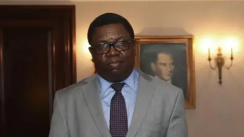 Anadolu Agency via Getty Images A bespectacled Joseph Malanji in a suit poses for a picture with a frame behind him 