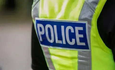 BBC The back view of a police officer wearing a high-vis yellow jacket with a sign on its back saying police in white letters on a blue background