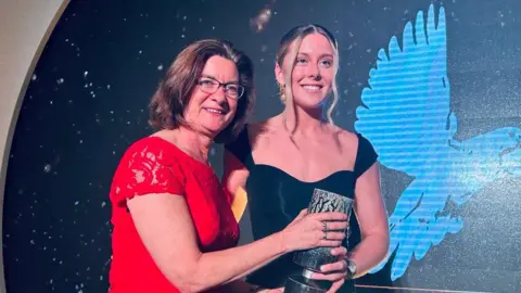 St David Award/X Eluned Morgan, who is wearing a red dress and glasses, is presenting the St David award to Emma Finucane who is wearing a black dress. They are standing in front of a dark background with a blue figure of a bird. 