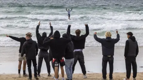 Surfing England Several highly-animated people on a beach look out at a surfer. Most of them are waving their arms. The surfer has his arms up in celebration.