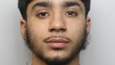 A young Asian man with thick dark hair, a thin moustache and straggly beard looks into the camera.
