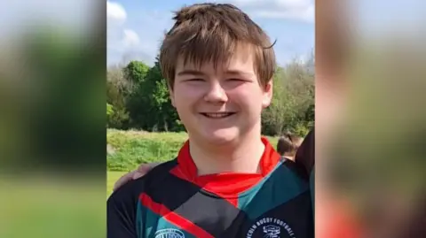 Lincoln Rugby Club A head and shoulders photo of a teenager with windswept brown hair with some of the fringe on his forehead. He is smiling and squinting at the camera and wearing a black, red and green rugby shirt. You can see trees behind him and someone has their arm around him.