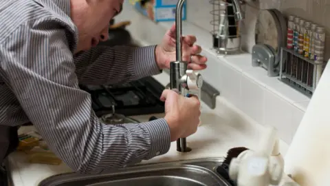 LDRS a man wearing a striped shirt is leaning over a chrome kitchen sink. He is holding the base of a chrome kitchen sink tap. He looks as if there is a problem and he is trying to repair it.