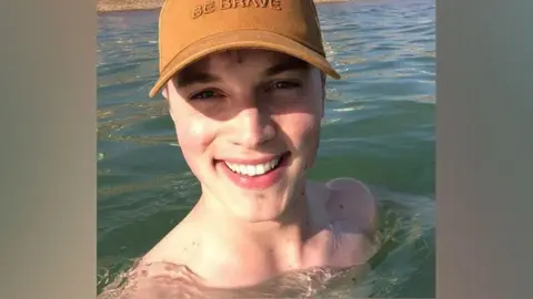 Family A photo of a man in the sea on a sunny day. There is water up to his shoulders and he is wearing a yellow baseball cap. He is smiling.