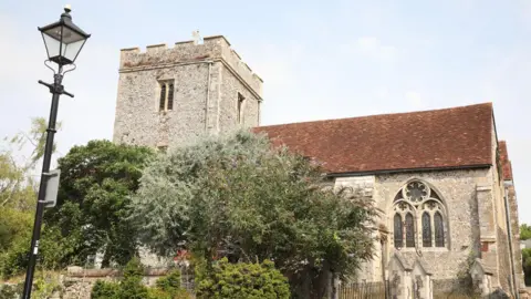 Ed Baker The church with its flint walls and red-tiled roof is bathed in sunshine with a gold weather vain on top of the bell tower.