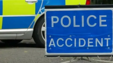 BBC A yellow and blue fluorescent police car which is parked on a road. Next to it is a blue sign which in white writing says "Police accident".