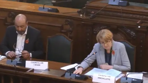 Bradford Council Susan Hinchliffe wears a grey jacket and sits in a large chair in a dark wood panelled room at Bradford Council meeting