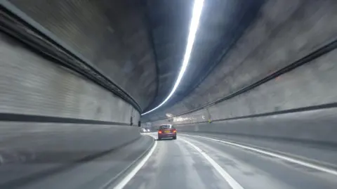 Getty Images A red car driving through the Rotherhithe tunnel. The tunnel has grey walls and curves to the left. Another car is approaching in the distance, in the opposite direction. 