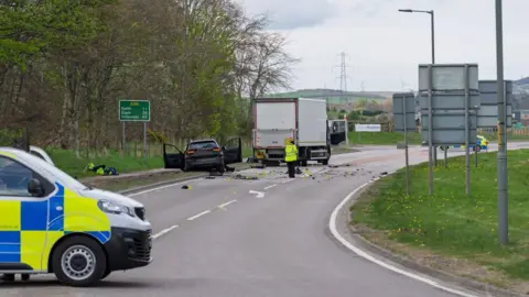 JasperImage Scene of crash, police vehicle in foreground, damaged black car and lorry beyond it.