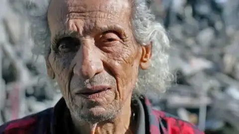 Alex Shaw/BBC A man, Fouad Hassan, stares off into the distance. Behind him is a twisted pile of concrete and metal rubble. Some of his family used to live where the rubble is now piled high.