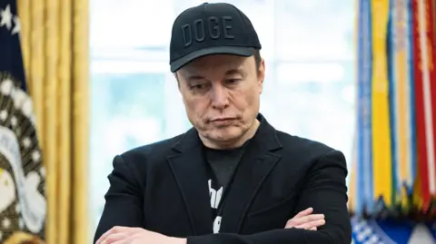 The Washington Post/Getty Images Elon Musk stands in the oval office in a black suit jacket, a black tshirt and a black cap with the letters DOGE stitched in. His arms are folded and he is looking downwards. In the background the flag with the President's standard can be seen to the right, it is out of focus.