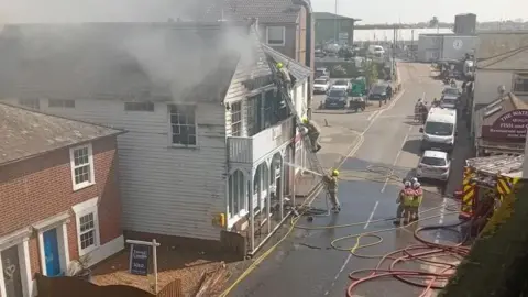 Martyn Bignell A white building on the left has white smoke coming out of the windows and the roof as there is a fire in the building.  On the road in front of the house are multiple firemen, and three of them are going up a ladder into the building.