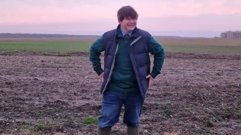 Submitted A pink sunset can be seen creating a hue across a photo. In the foreground a teenager with brown hair and a fringe can be seen laughing and looking to the side. He is wearing a navy waistcoat, blue fleece with a school tie and shirt underneath. He also has jeans and wellies on and is standing in a field.