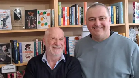 Colin Wade Scheme founders Colin Wade and Laury Conn smiling for the camera in front of a book shelf in the Mostly Books shop in Abingdon.