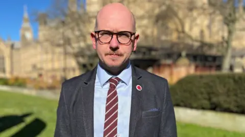 BBC A bald man with glasses outside the Westminster Parliament on a sunny day. He is looking at the camera and is wearing a grey suit, blue shirt and maroon and grey striped tie. 